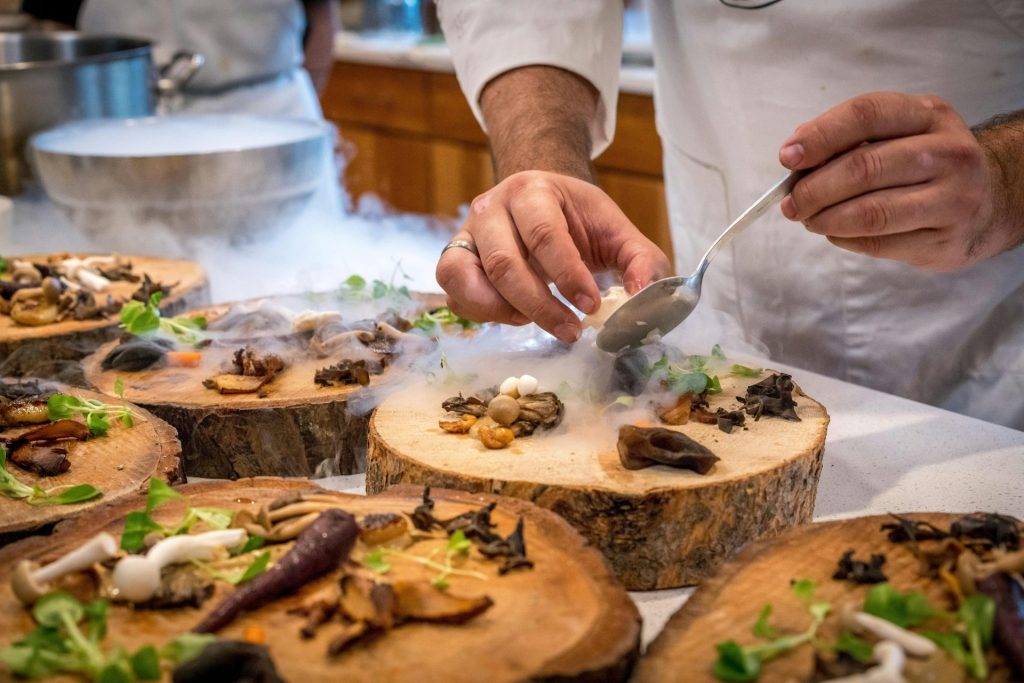 Ein Koch präsentiert kunstvoll ein Gourmetgericht mit Pilzen und Gemüse auf Holzplatten.